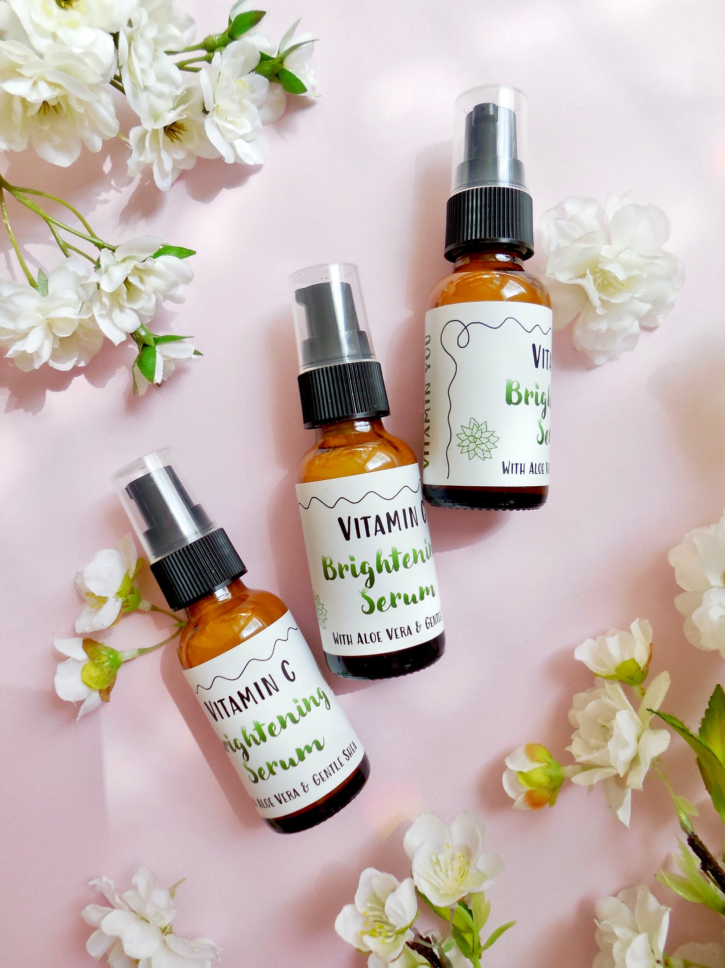 Three bright bottles of the Vitamin C Serum on a pink background surrounded by flowers