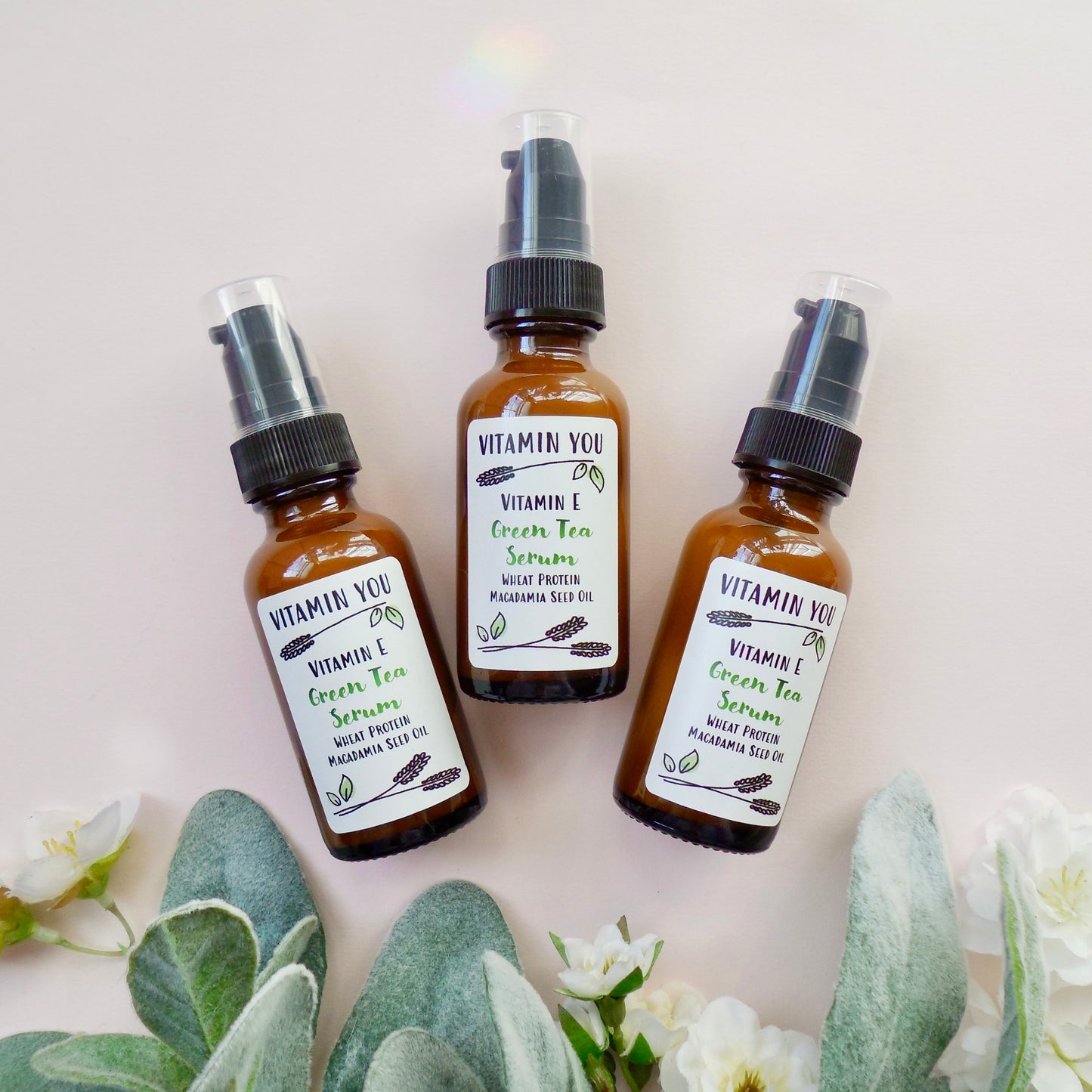 Three bottles of the Green Tea Serum next to flowers and leaves on a light pink background