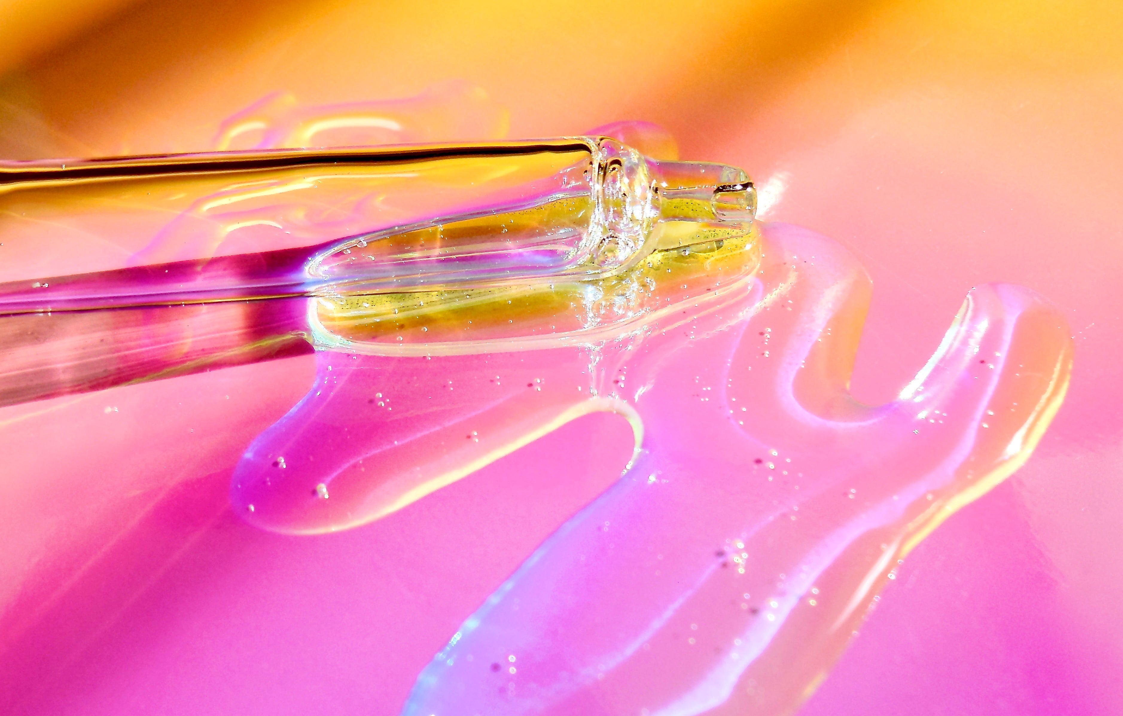 Close up image of a serum dropper and clear aloe vera serum in a sunset gradient of pinks and yellows. 