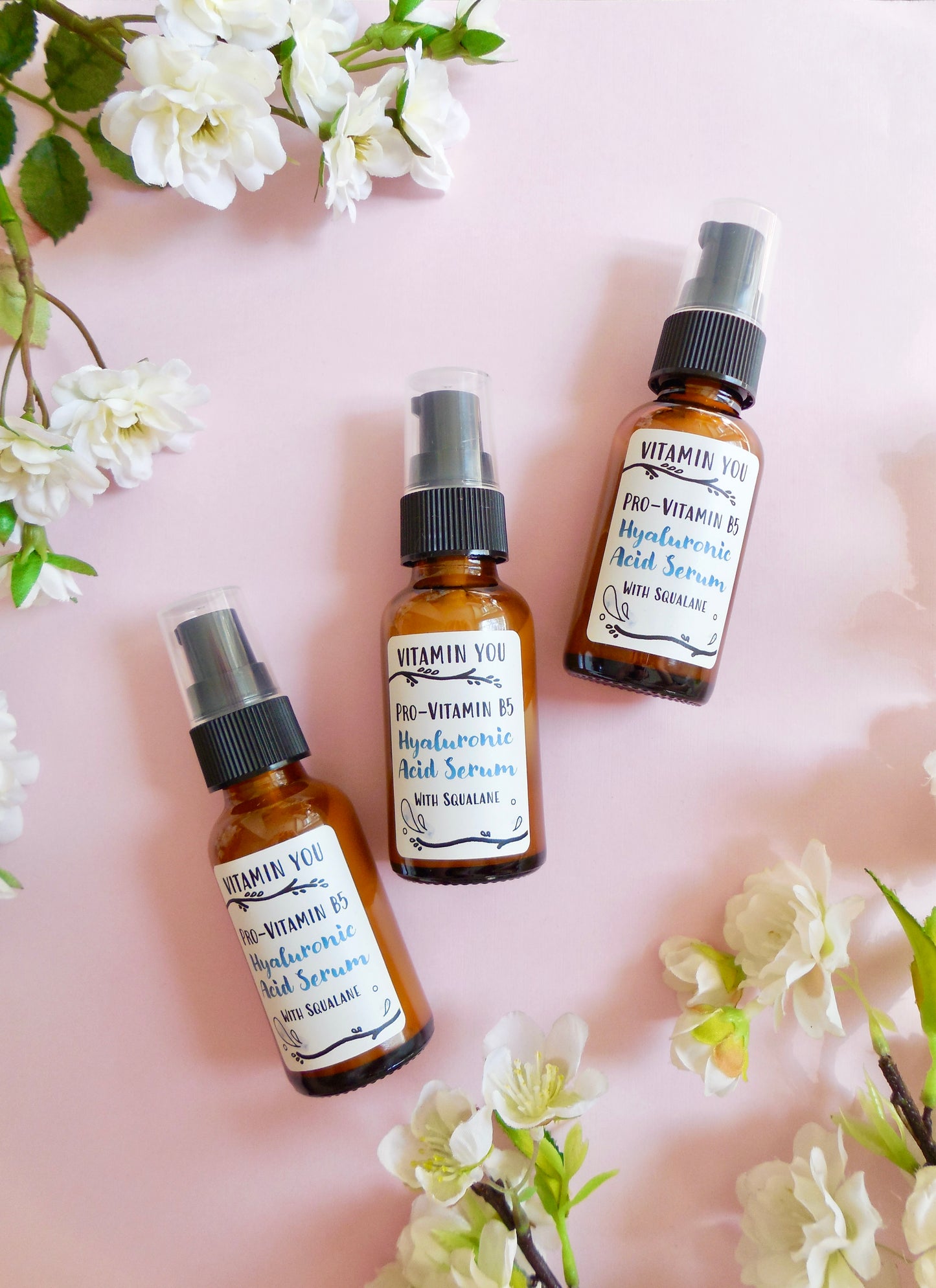 Bottles of Hyaluronic Acid Serum on a pink background surrounded by flowers