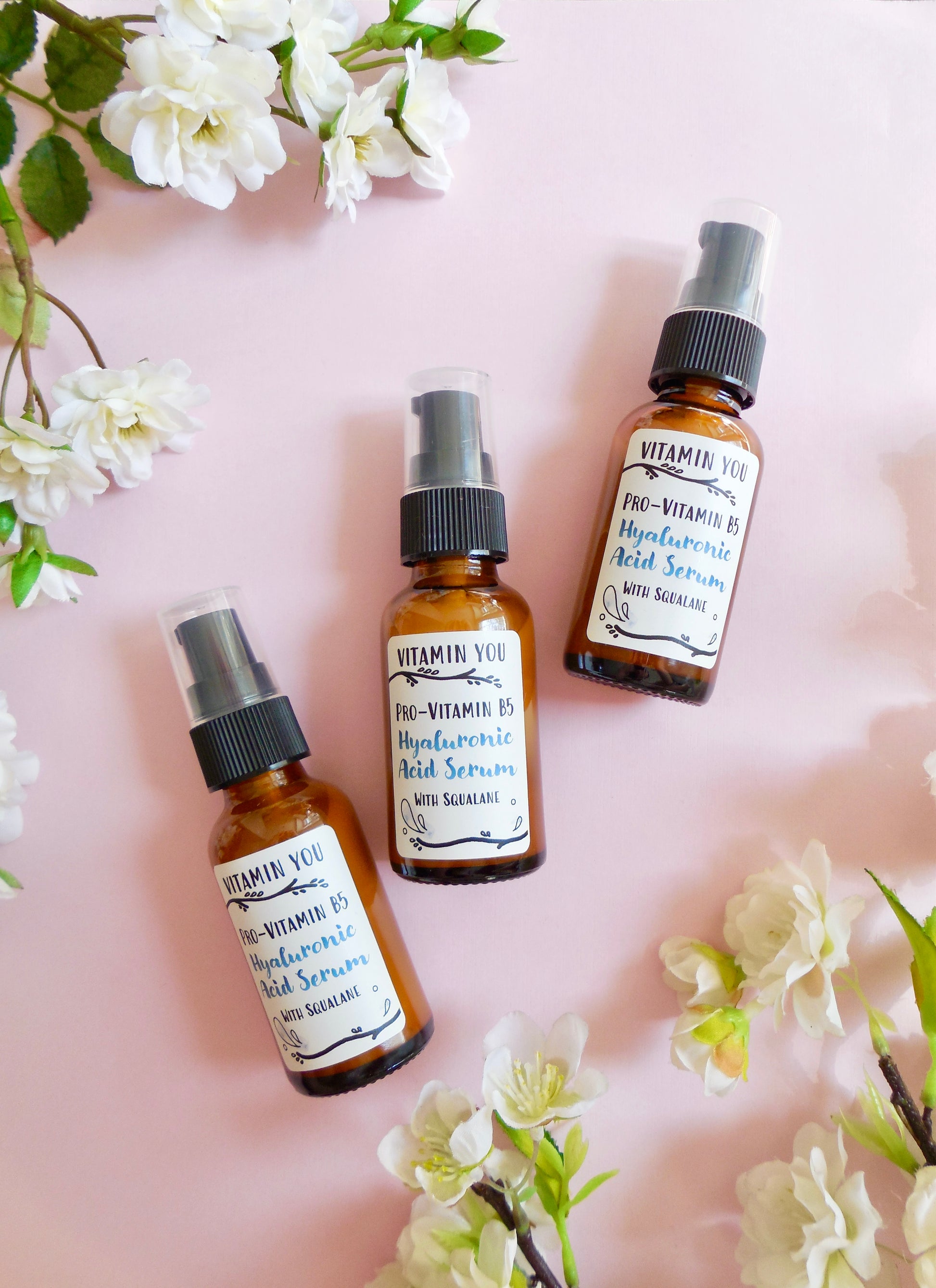 Bottles of Hyaluronic Acid Serum on a pink background surrounded by flowers