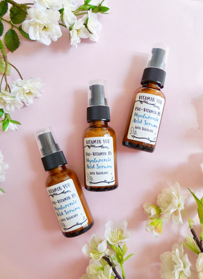 Bottles of Hyaluronic Acid Serum on a pink background surrounded by flowers