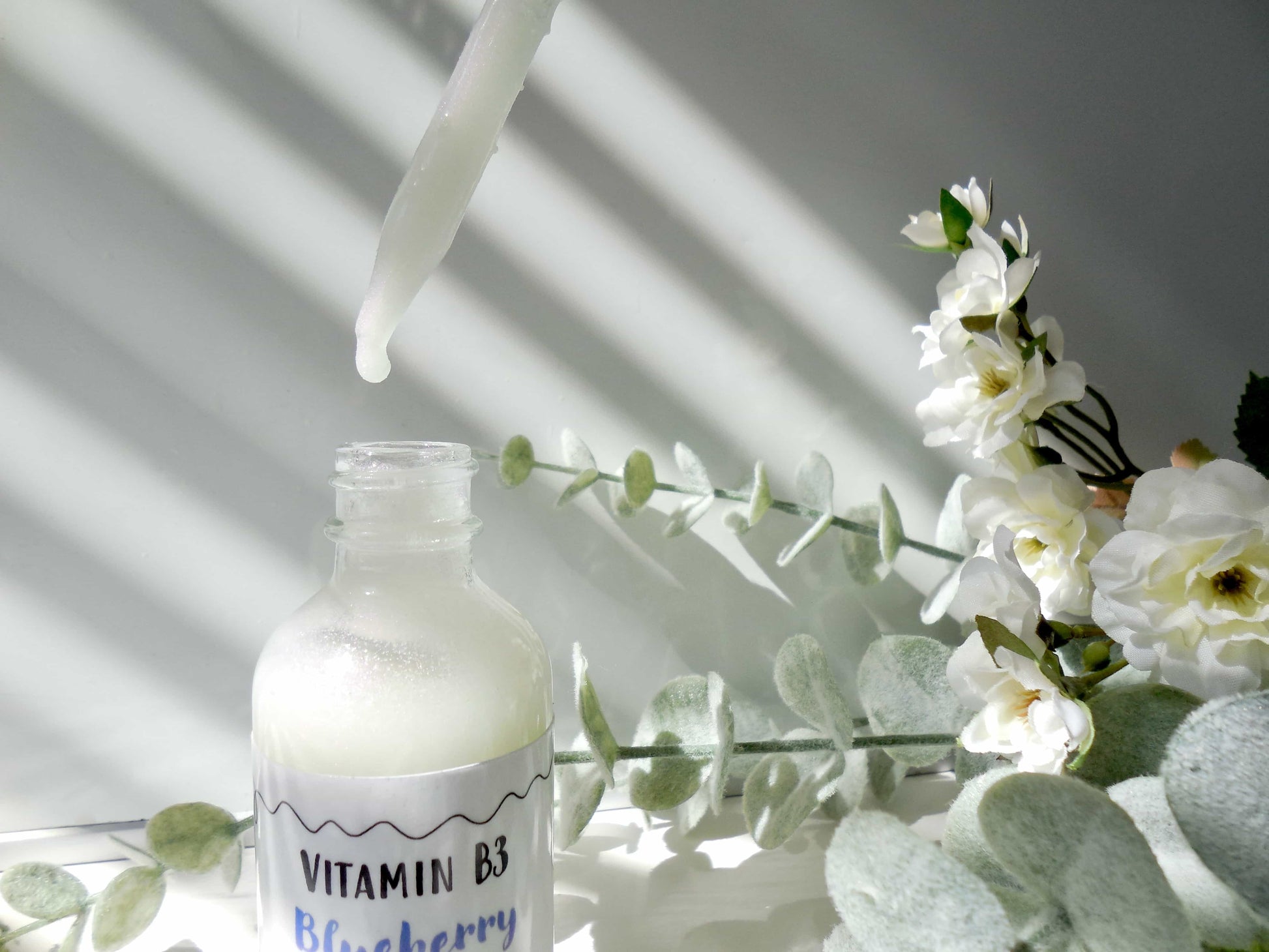 Image of the white moisturizer being used with the dropper tool next to eucalyptus and flowers. 